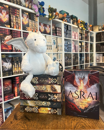 Das Produktbild zeigt den weißen Plüsch-Drachen Manila (aus der Asrai-Buchreihe von Liane Mars) von vorn auf einem Stapel Asrai-Bücher sitzend vor Bücherregalen. Daneben sieht man frontal das Buch "Asrai - Das Herz der Drachen".